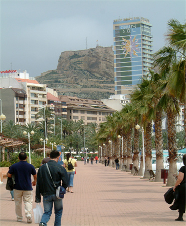 Bild på människor som går nedför en gata. Gatan kantas av palmer. I bakgrunden syns en borg högt uppe på ett berg.